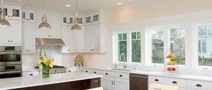 Bright, modern kitchen with white cabinets, stainless steel appliances, and a large center island with pendant lighting. The space features multiple windows along one wall, letting in natural light, and a countertop decorated with a vase of flowers and a fruit stand.