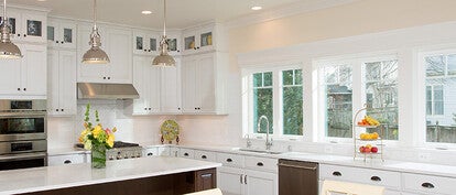 Bright, modern kitchen with white cabinets, stainless steel appliances, and a large center island with pendant lighting. The space features multiple windows along one wall, letting in natural light, and a countertop decorated with a vase of flowers and a fruit stand.