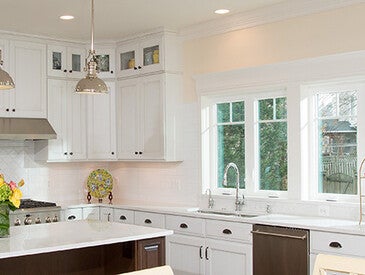 Bright, modern kitchen with white cabinets, stainless steel appliances, and a large center island with pendant lighting. The space features multiple windows along one wall, letting in natural light, and a countertop decorated with a vase of flowers and a fruit stand.