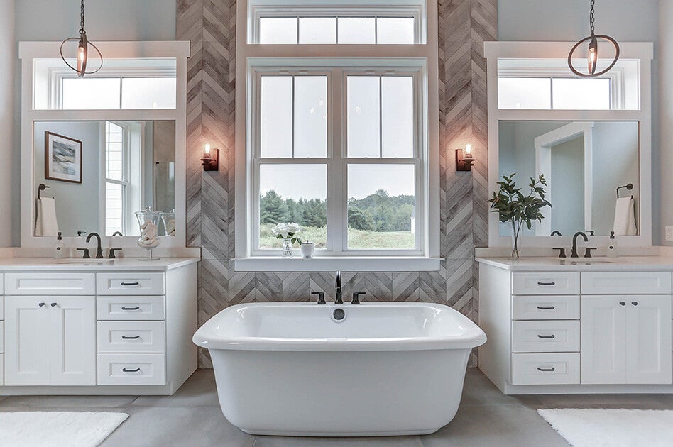 Elegant bathroom featuring a freestanding white bathtub centered between two vanities with white cabinetry and large mirrors. The space includes modern light fixtures, herringbone tile accents, and a large window providing natural light and a scenic outdoor view.