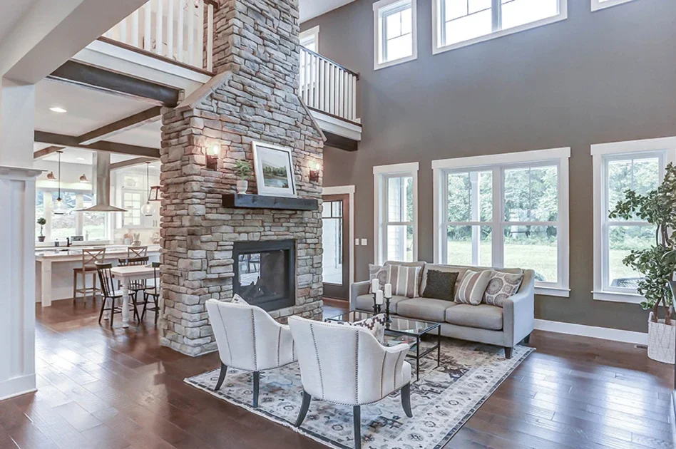 Bright and spacious living room with tall ceilings, large windows, and a floor-to-ceiling stone fireplace. The room features a gray sofa, two accent chairs, and a patterned rug, with an open view into a modern kitchen and dining area.