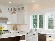 Bright, modern kitchen with white cabinets, stainless steel appliances, and a large center island with pendant lighting. The space features multiple windows along one wall, letting in natural light, and a countertop decorated with a vase of flowers and a fruit stand.