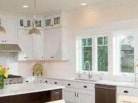 Bright, modern kitchen with white cabinets, stainless steel appliances, and a large center island with pendant lighting. The space features multiple windows along one wall, letting in natural light, and a countertop decorated with a vase of flowers and a fruit stand.