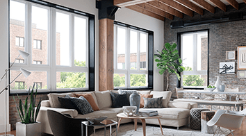 Modern Loft apartment living room featuring several tall windows. The room has exposed rafter ceilings, a brick wall and a large l shaped sectional.