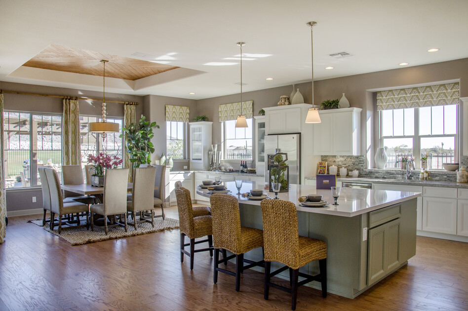 A spacious open-concept kitchen and dining area with modern decor. The kitchen features white cabinetry, stainless steel appliances, and a large island with woven barstools. The adjoining dining area has an elegant table set for eight beneath a pendant light, with large windows bringing in natural light and views of the outdoors.