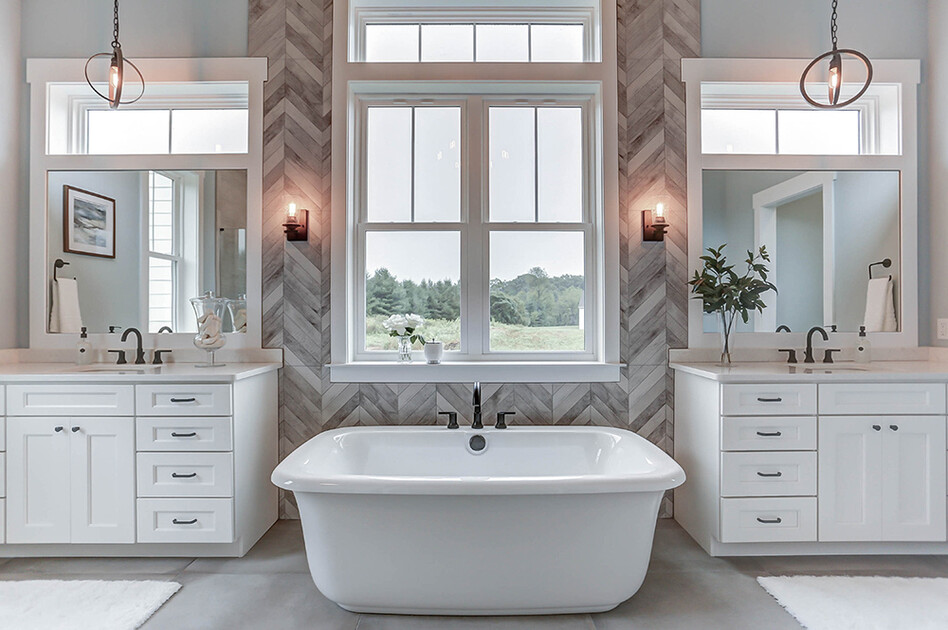 Hummelstown Case Study Bathroom Elegant bathroom featuring a freestanding white bathtub centered between two vanities with white cabinetry and large mirrors. The space includes modern light fixtures, herringbone tile accents, and a large window providing natural light and a scenic outdoor view.
